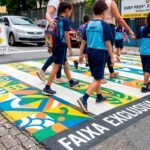 Crianças e pais atravessando rua sinalizada com urbanismo tático e faixa de pedestres elevada em frente a escola municipal